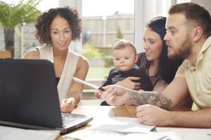 a young husband and wife with their baby sit and chat to a woman in their dining room . They are all referring to a laptop in front of them and various paperwork is dotted about the table.