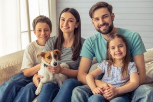 Beautiful young parents, their cute little daughter and son are looking at camera and smiling, sitting with their cute dog on sofa at home