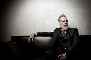 Portrait of adult businessman wearing trendy suit and sitting modern studio on leather sofa against the empty wall.